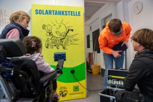 Ein E-Rolli und Tablets werden an der Solartankstelle geladen, mehrere Schüler:innen stehen daneben