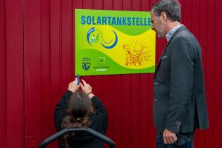 Eine Schülerin schraubt ein Schild mit dem Tital "Solartankstelle" an eine rote Wand, ein Lehrer steht daneben