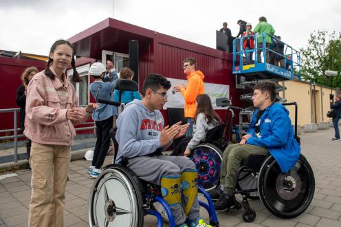 Mehrere Schüler:innen, ein paar davon im Rollstuhl, stehen auf dem Schulhof vor einem roten Container
