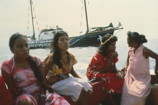 Frauen sitzen in einem Boot, im Hintergrund die Rainbow Warrior