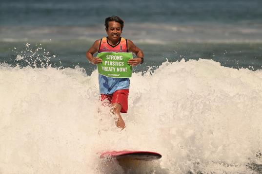 Surfer auf dem Meer hält ein Banner "Strong Plastics Treaty Now!"