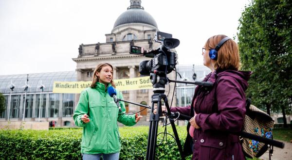 Greenpeace-Expertin vor einer Kamera, im Hintergrund die Münchener Staatskanzlei und ein Banner “Windkraft statt heißer Luft, Herr Söder“.