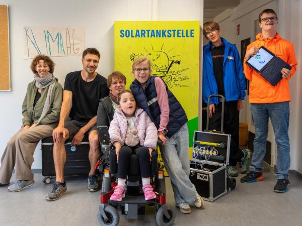 Drei Erwachsene und vier Schüler:innen, eine davon im Rollstuhl, stehen vor der gelben Solartankstelle